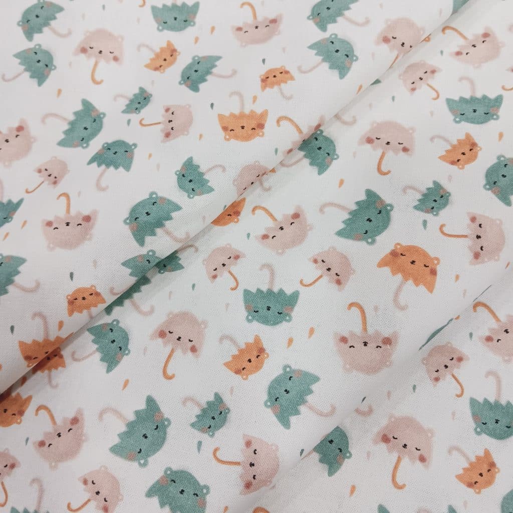 Tela de algodón con estampado de paraguas con caritas adorables en tonos pastel: verde menta, rosa y mostaza, sobre fondo blanco. Estilo suave, infantil y alegre.