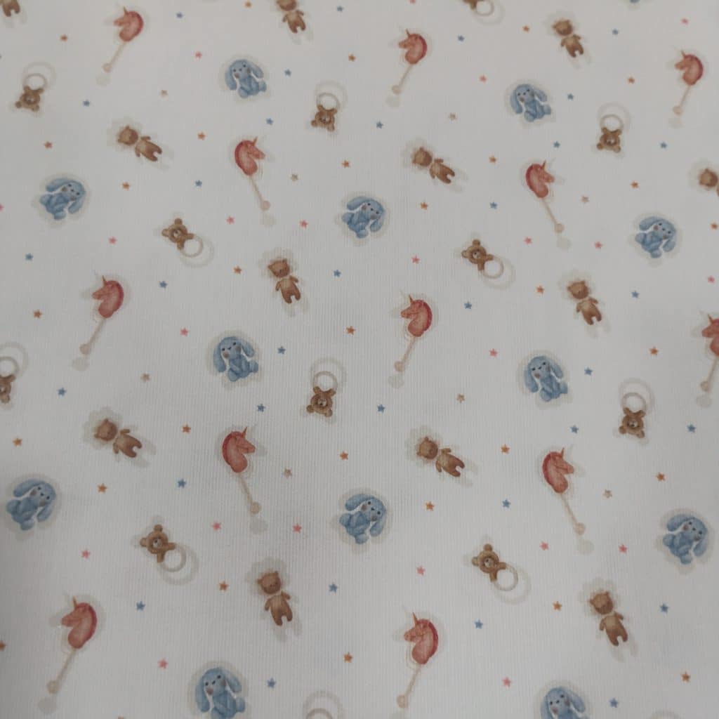 Piqué de algodón 100% en fondo blanco tenemos un estampado de juguetitos infantiles como conejito de tela azul, sonajero en forma de osito y caballito de madera
