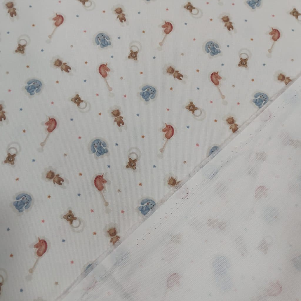 Piqué de algodón 100% en fondo blanco tenemos un estampado de juguetitos infantiles como conejito de tela azul, sonajero en forma de osito y caballito de madera