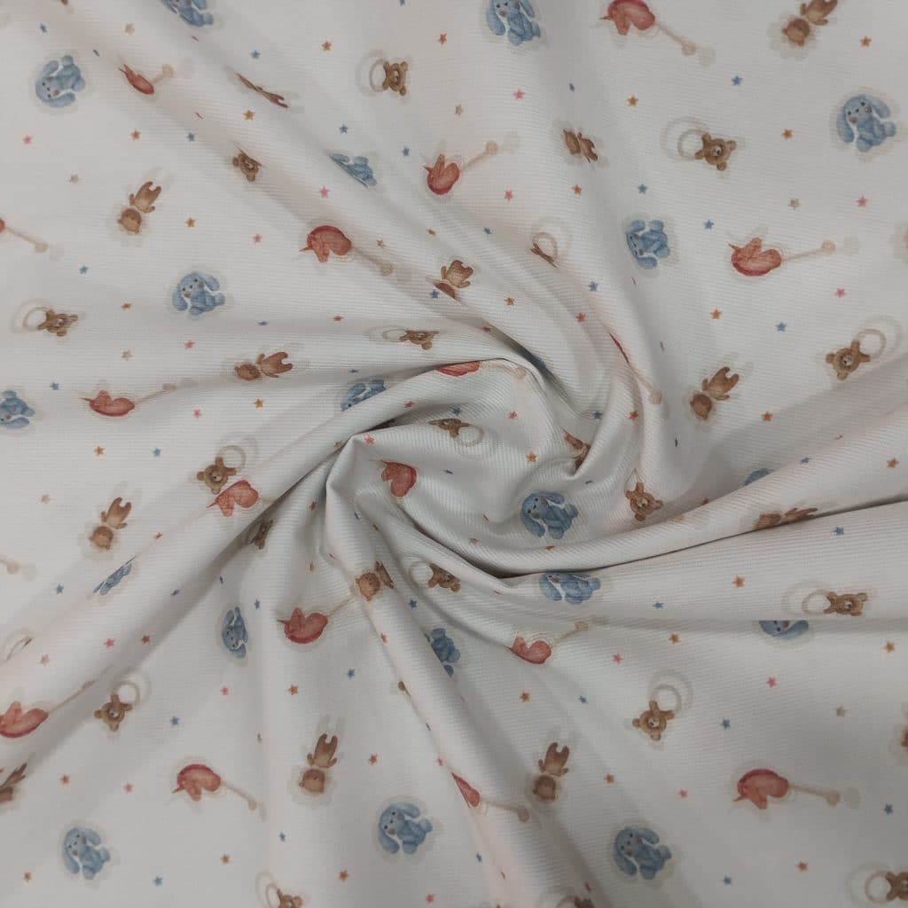 Piqué de algodón 100% en fondo blanco tenemos un estampado de juguetitos infantiles como conejito de tela azul, sonajero en forma de osito y caballito de madera