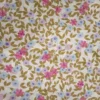 Tela de algodón blanca con estampado de pequeñas flores rosas, azules y lilas, acompañadas de ramas y hojas mostaza sobre fondo blanco.