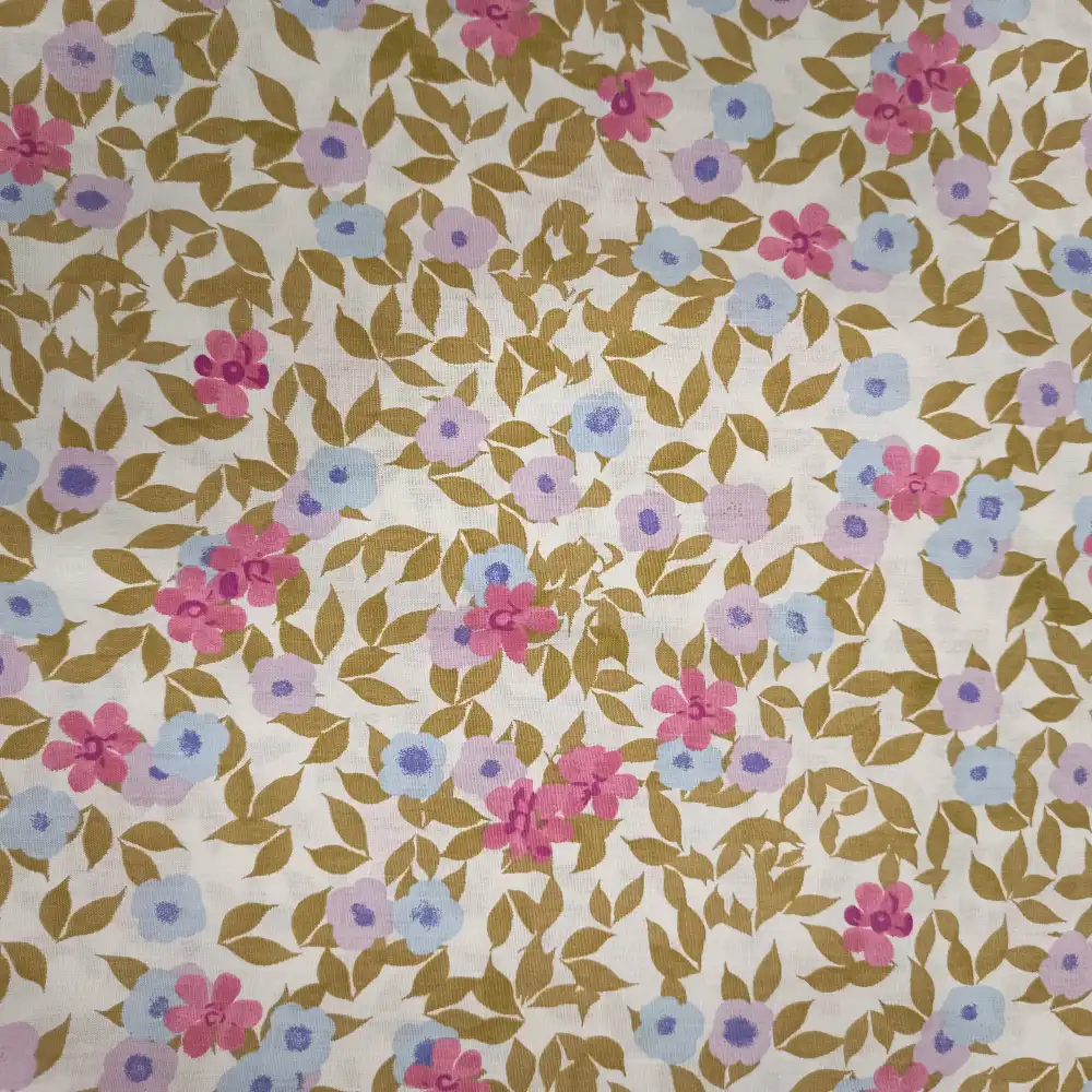 Tela de algodón blanca con estampado de pequeñas flores rosas, azules y lilas, acompañadas de ramas y hojas mostaza sobre fondo blanco.
