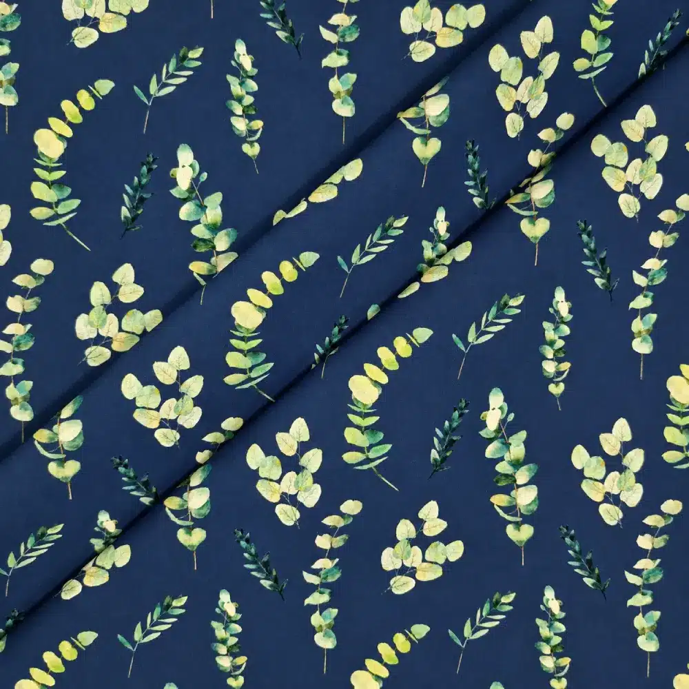 Tela de algodón azul oscuro con estampado de ramas y hojas de eucalipto en diferentes tonos verdes, amarillos suaves y crema, distribuidas sobre el fondo.