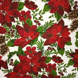 Tela de mantelería navideña blanca con flores de Pascua grandes en tonos rojos y burdeos, piñas marrones, bayas rojas y ramas verdes, dispuesta con pliegues diagonales.