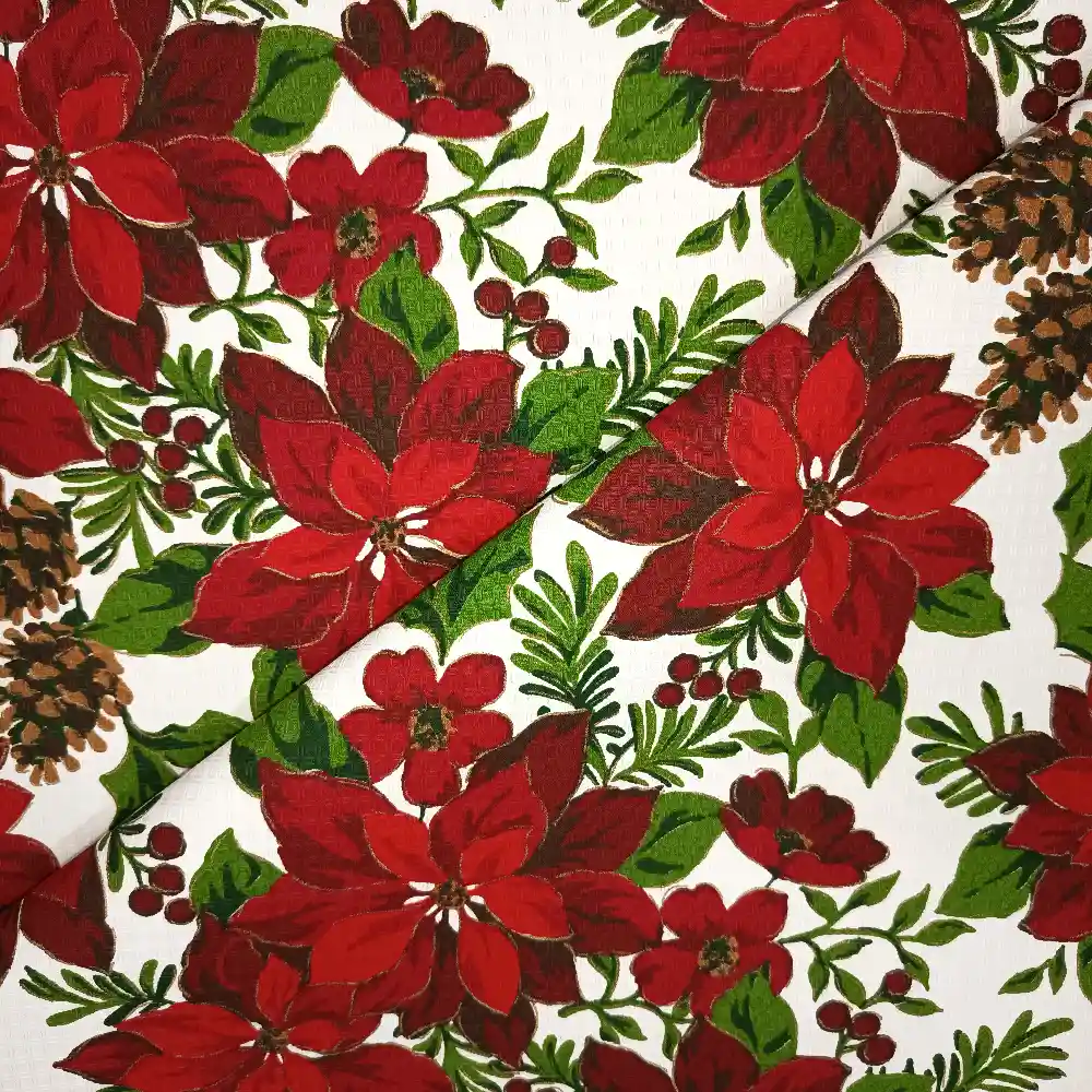 Tela de mantelería navideña blanca con flores de Pascua grandes en tonos rojos y burdeos, piñas marrones, bayas rojas y ramas verdes, dispuesta con pliegues diagonales.