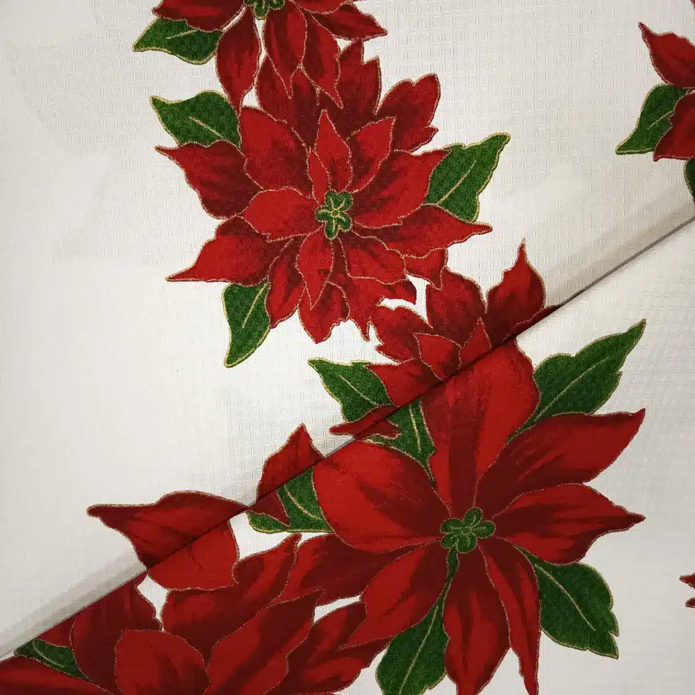 Tela de mantelería navideña blanca con grandes flores de Pascua rojas y burdeos, hojas verdes y finos contornos dorados, sobre fondo blanco con textura.